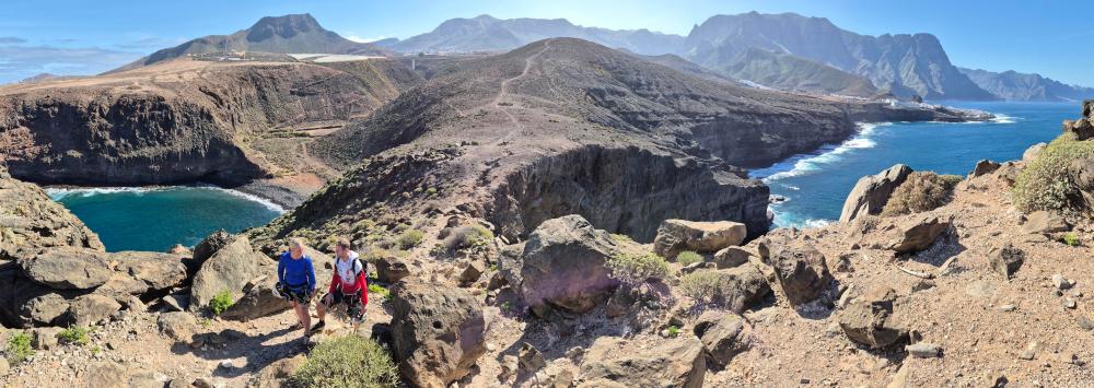 Two hikers on a rocky cliff overlooking a coastal landscape with mountains and ocean.