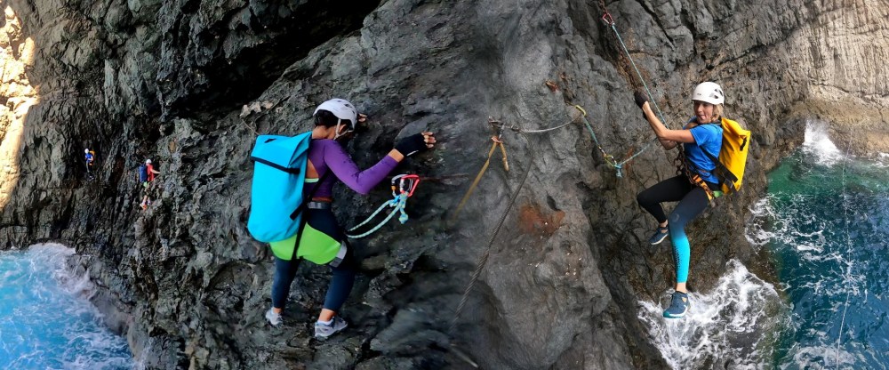 Two climbers on a rocky coastal cliff with safety gear above ocean waves.