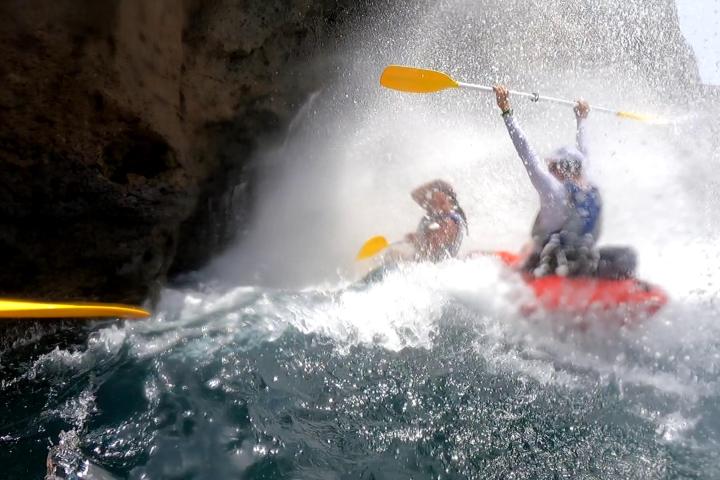 Two people kayaking through rough waves near a rocky cliff.