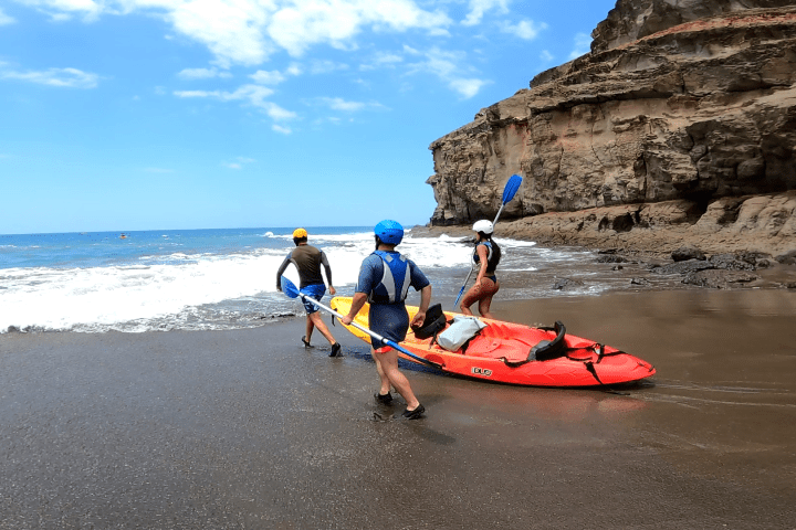 Three people prepare a red kayak on a sandy beach by a rocky cliff.