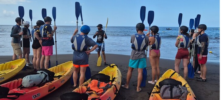 Group of people with kayaks and paddles on a beach, receiving instructions from a guide.