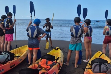 Group of people with kayaks and paddles on a beach, receiving instructions from a guide.