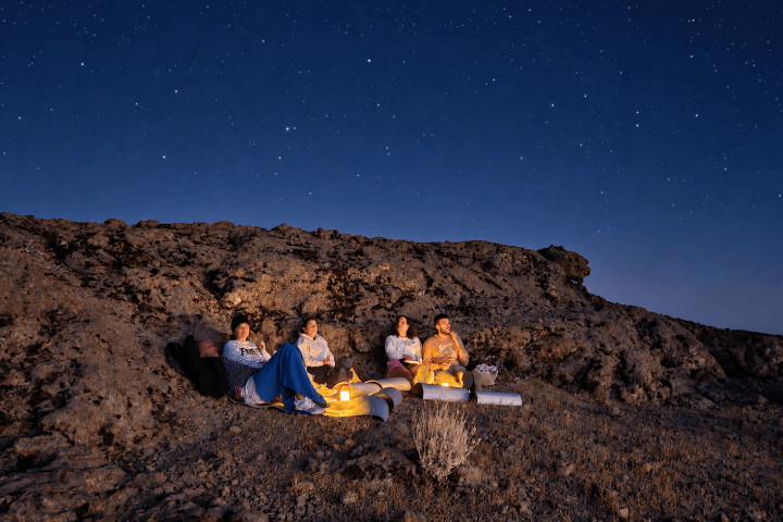 Four people sitting on a rocky hill under a starry night sky.