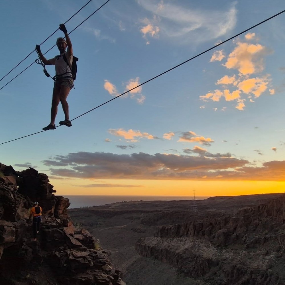 Person crossing a canyon on a zip line at sunset, wearing safety gear.