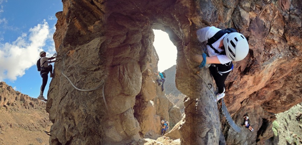 People climb rocky mountain trail with safety gear, blue sky visible.