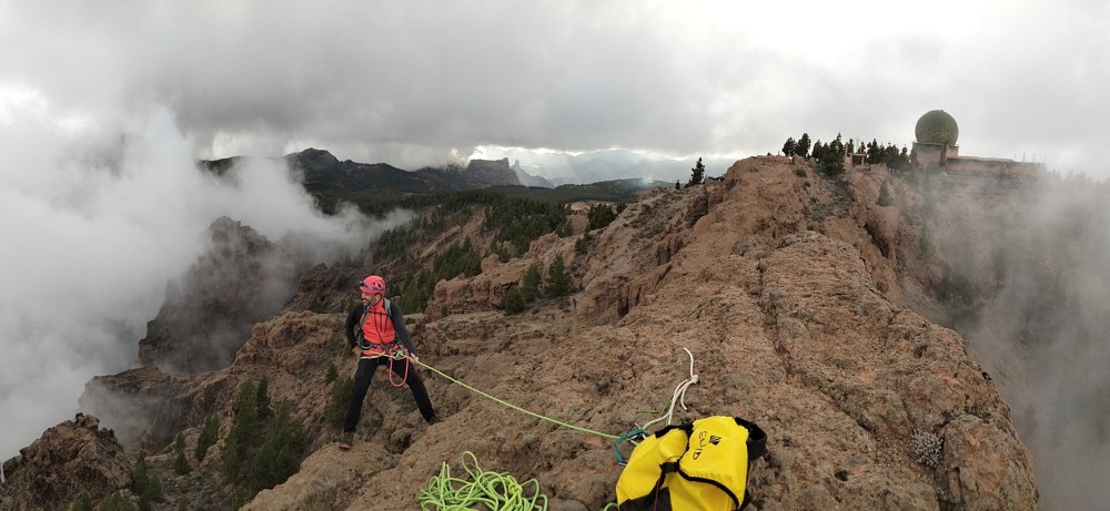 Climber on rocky ridge with foggy mountain view and yellow bag nearby. Radar dome in background.