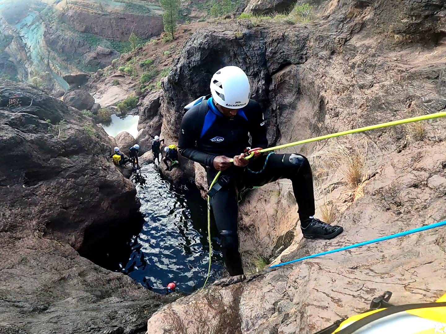 Person rappelling down a rocky canyon with others in wetsuits nearby.