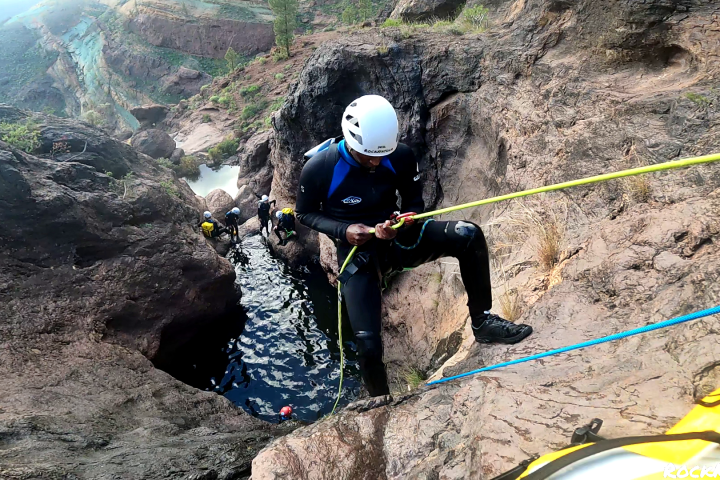 Person rappelling down a rocky canyon with others in wetsuits nearby.