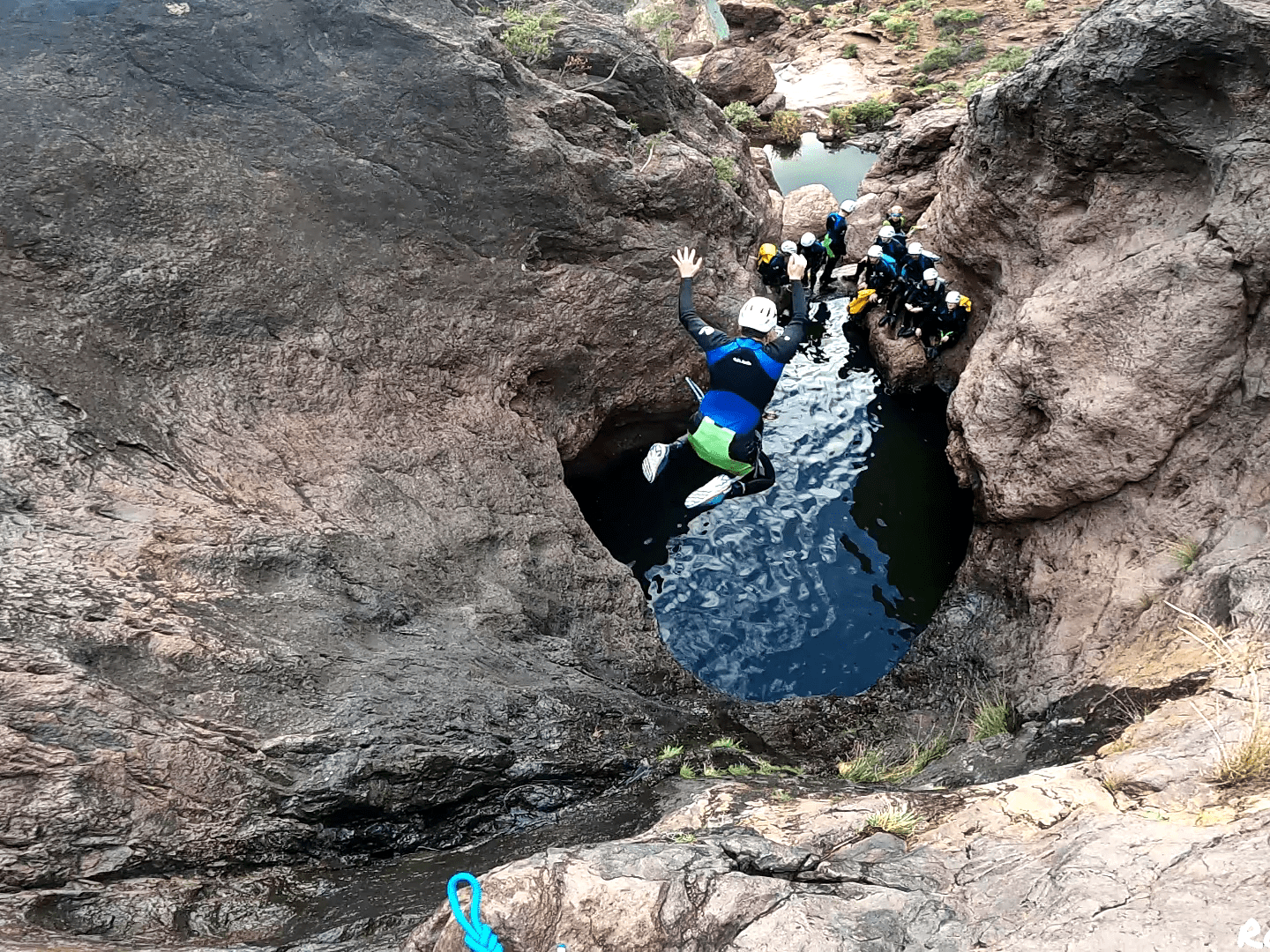 Person in helmet jumps into narrow water pool surrounded by rocky terrain, with onlookers nearby.