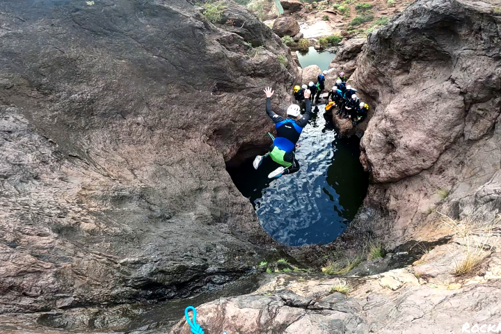 Person in helmet jumps into narrow water pool surrounded by rocky terrain, with onlookers nearby.