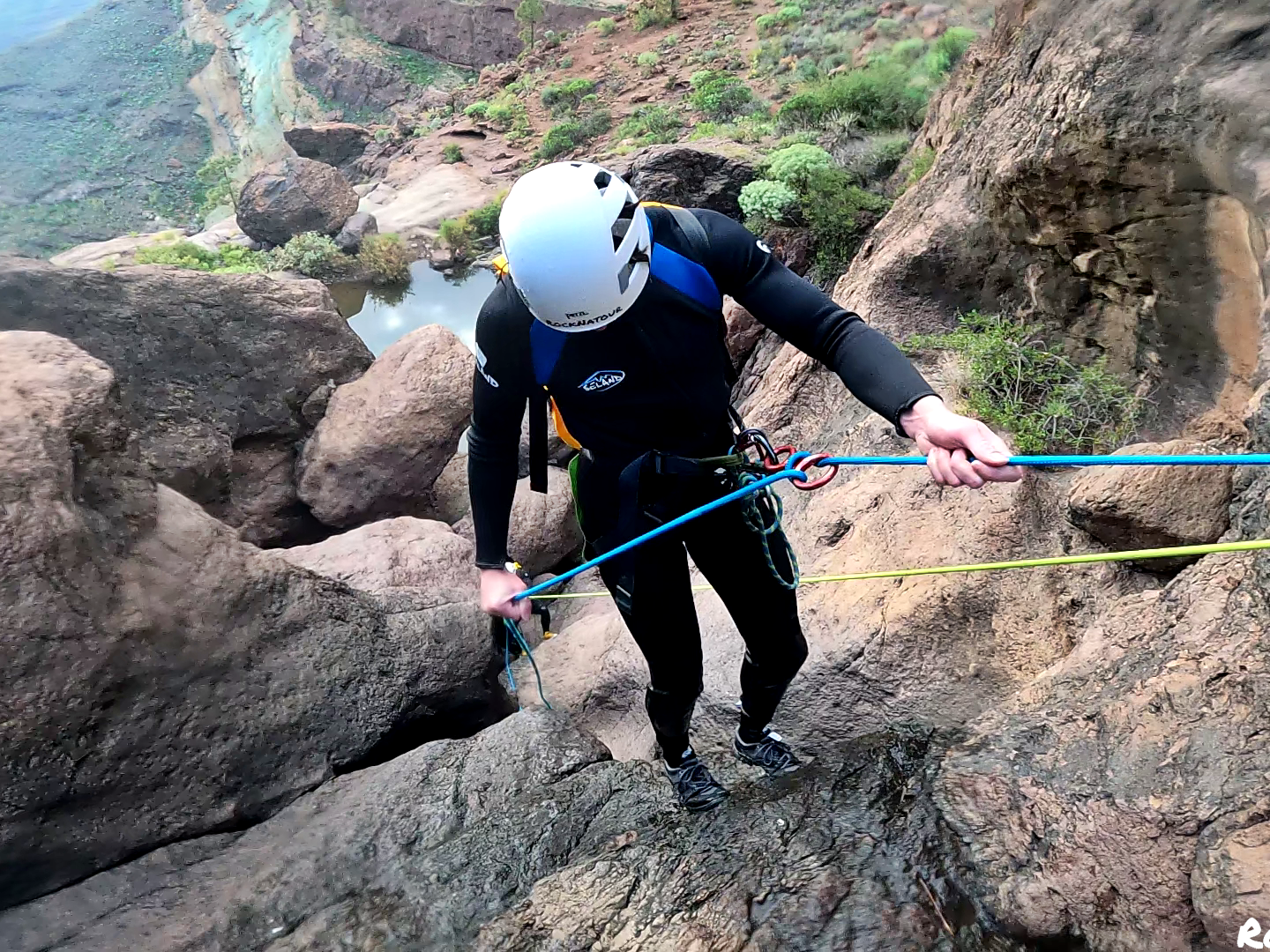 Person in helmet rappelling down a rocky slope near a river in a canyon landscape.