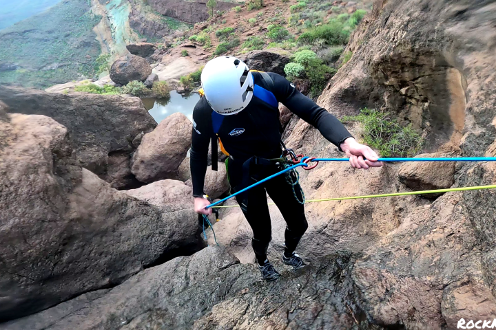 Person in helmet rappelling down a rocky slope near a river in a canyon landscape.