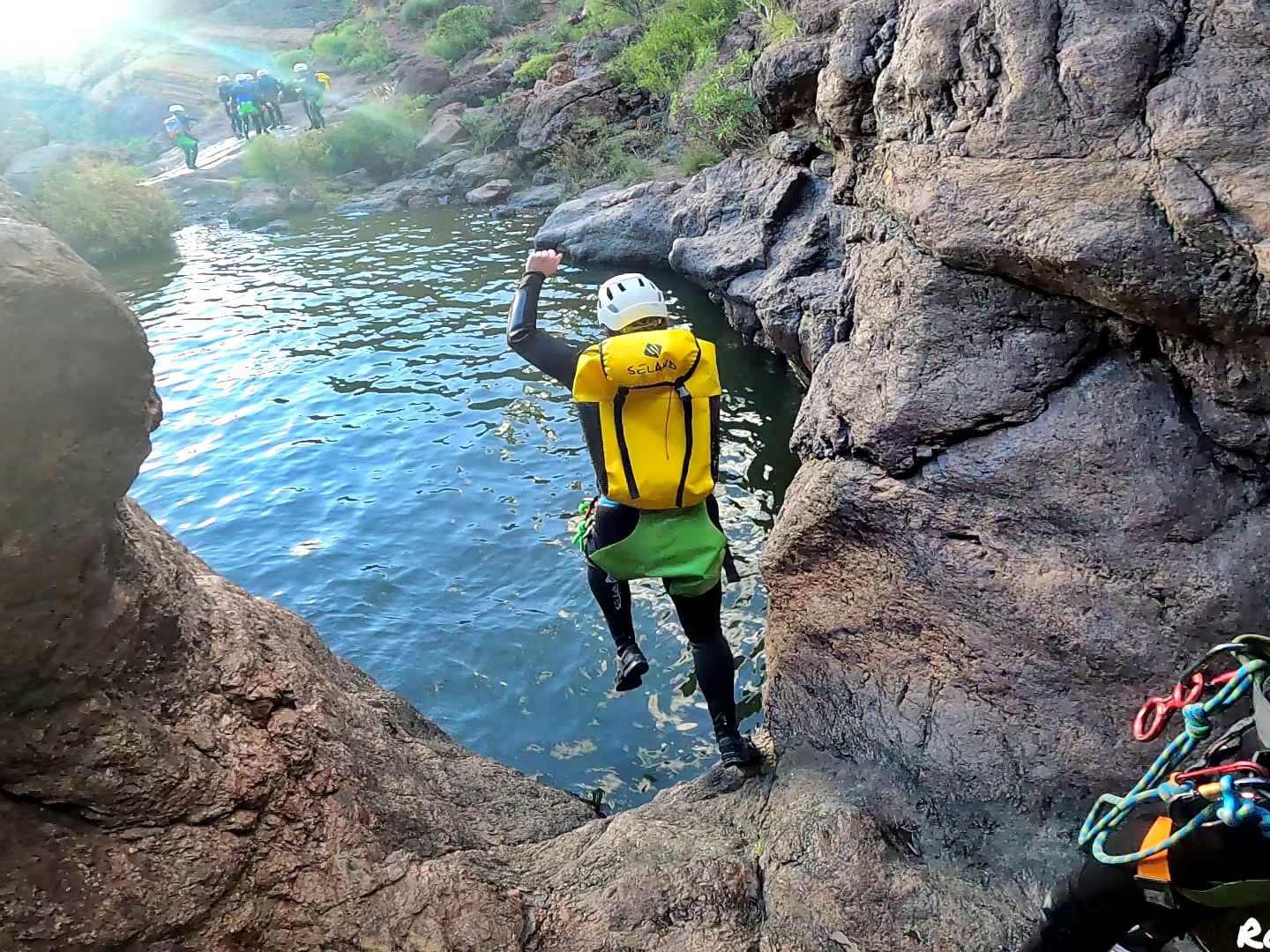 Person in helmet jumps from rocky ledge into river, others watch.