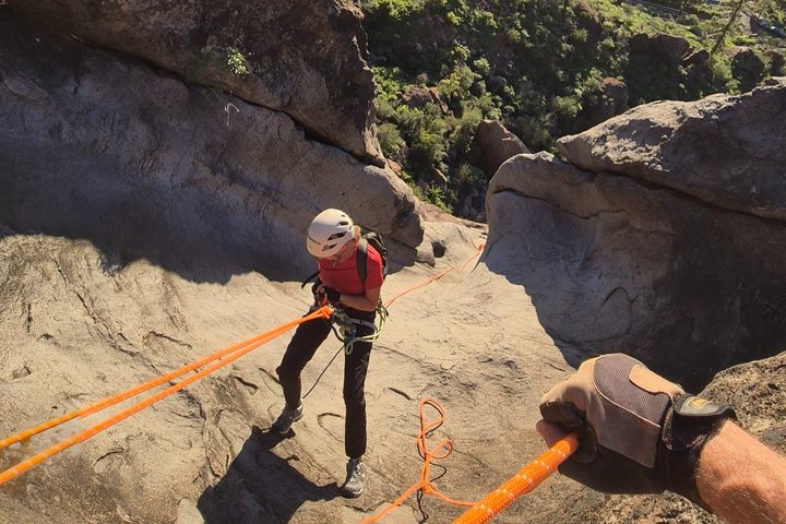 Person wearing helmet rappelling down rocky slope using orange ropes.