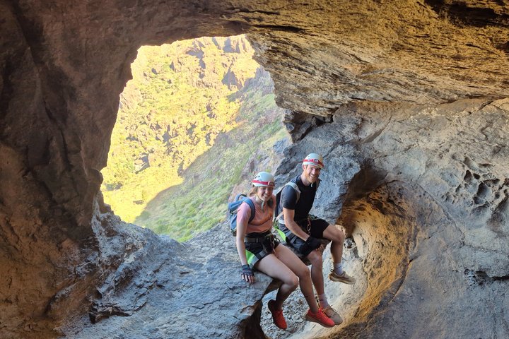 Two hikers sit on a rocky ledge inside a cave opening with a view of greenery outside.