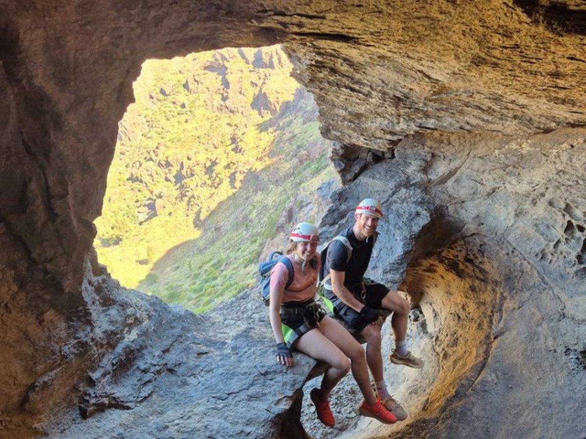 Two hikers sit on a rocky ledge inside a cave opening with a view of greenery outside.