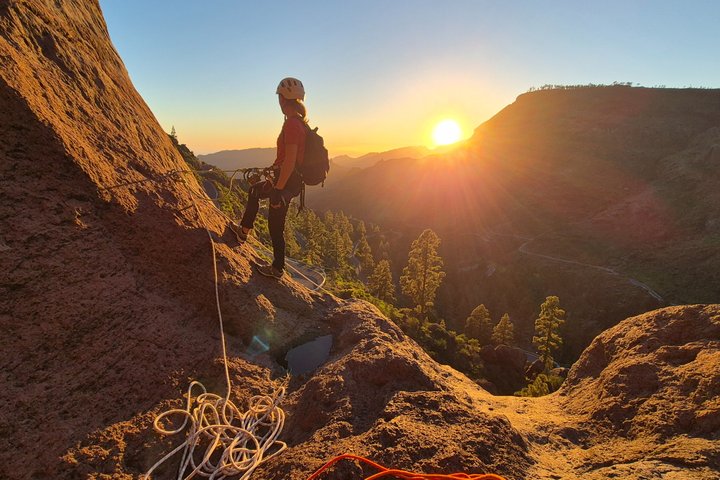 Climber on rocky slope at sunset with climbing gear and distant hills.