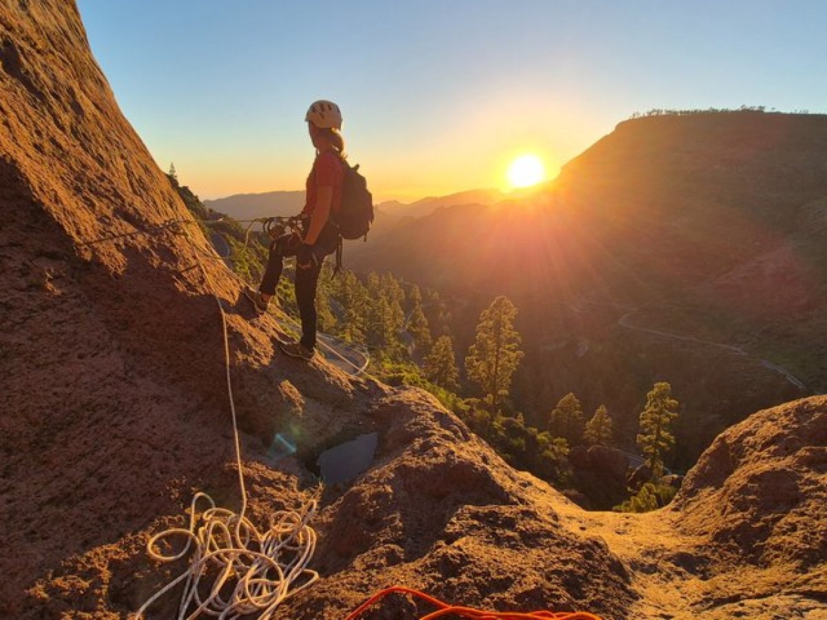 Climber on rocky slope at sunset with climbing gear and distant hills.